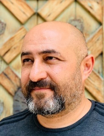 Fatih Başbuğ'un portresi: sakallı, gri tişörtlü, ahşap geometrik bir kapının önünde yan bakıyor.